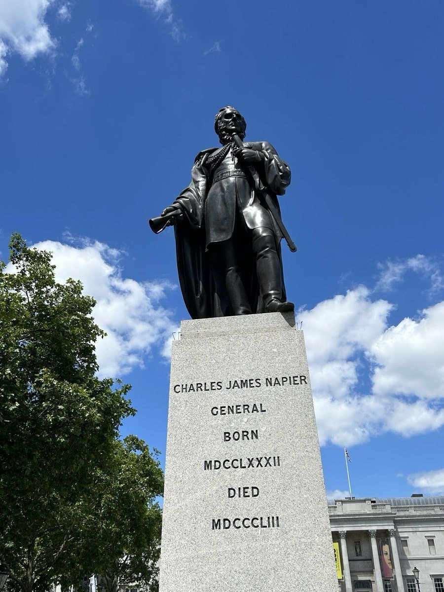 Bronze statue of Sir Charles James Napier in military dress on a granite pedestal, against a blue sky with cumulus clouds, Trafalgar Square, London.