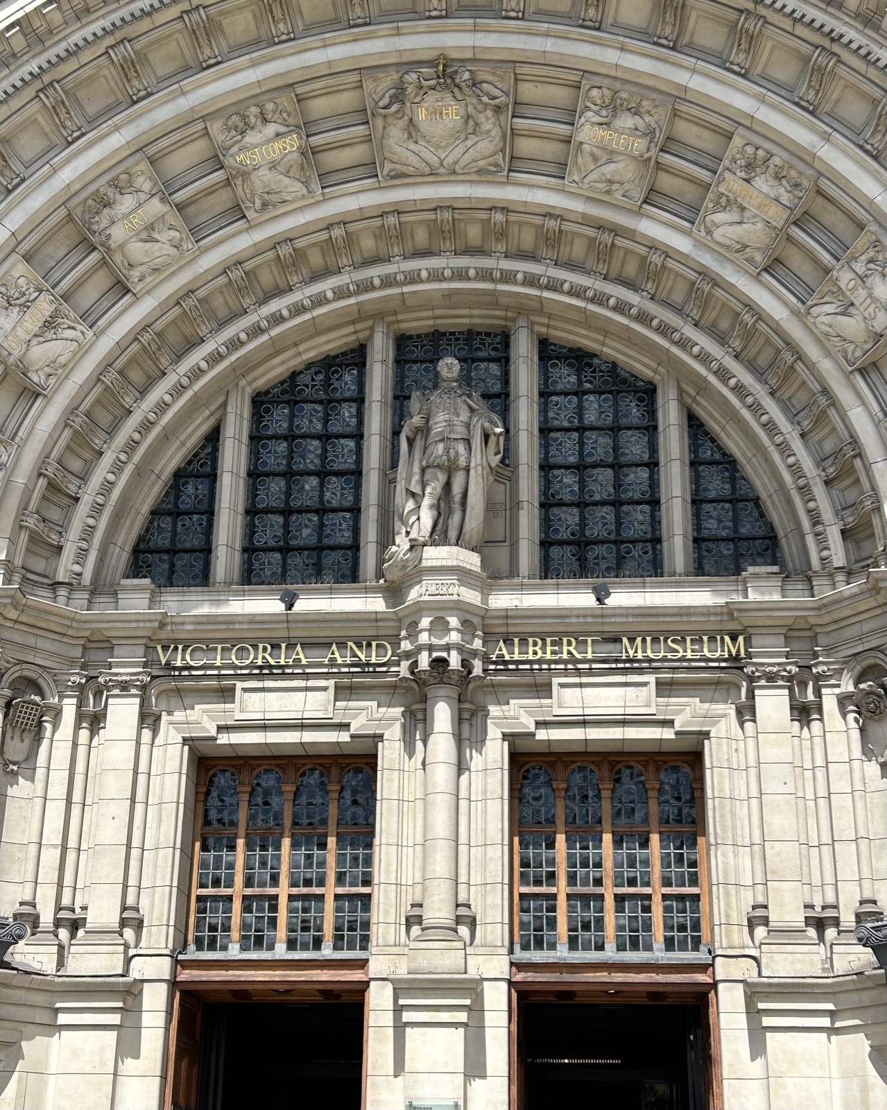 The arched stone entrance to the Victoria and Albert Museum in South Kensington, London, with the words VICTORIA AND ALBERT MUSEUM carved into the entablature and a sculpted figure under the arch.